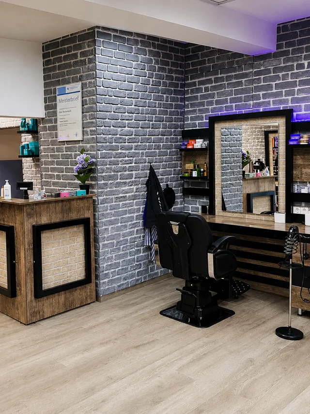 Blick in den Salon: Holztresen, graue Steinwand und schwarzer Barberstuhl vor dem Spiegelplatz, daneben der gerahmte Meisterbrief